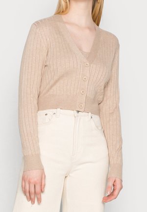 Kvinde iført en beige cropped ribstrikket cardigan med fire knapper, kombineret med højtaljede off-white bukser, hænderne afslappet langs siderne.