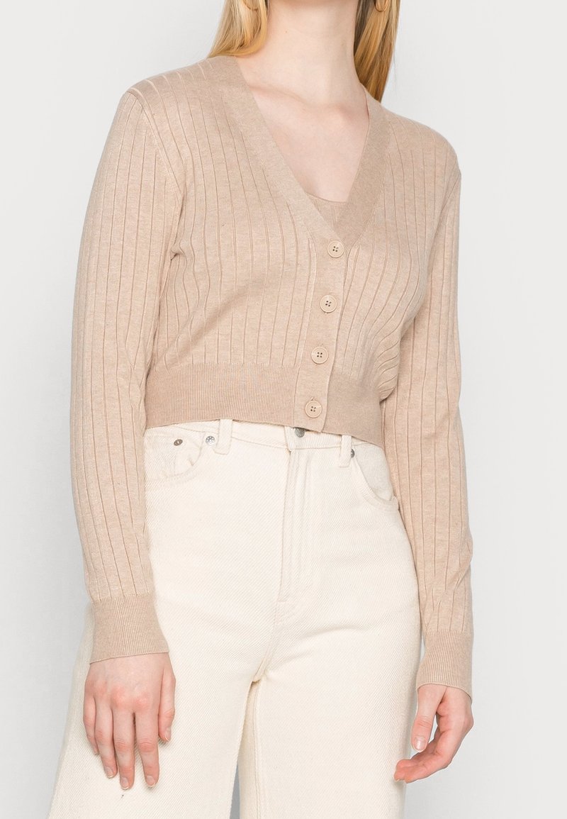 Femme portant un cardigan court côtelé beige à quatre boutons, associé à un pantalon taille haute blanc cassé, les mains détendues le long du corps.