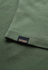 Groen ribbeltextiel met een gevouwen rand, met een marineblauwe label waarop "SUPERDRY" in goud staat. Zachte textuur en een consistent patroon overal.
