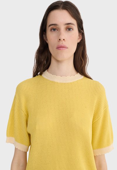 Jeune femme aux longs cheveux bruns portant un pull court à manches courtes jaune côtelé avec un col et des bords de manches crème festonnés.