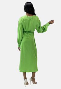 Groen, lang mouwen jurk met een getailleerde taille en een vloeiende rok. Gladde textuur en een ontspannen pasvorm, eindigend onder de knie. Gecombineerd met witte hakken.