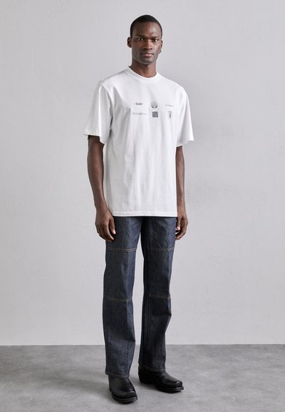 Weißes Baumwoll-T-Shirt mit Grafikdruck, kombiniert mit dunkelblauen Jeans aus Denim mit sichtbaren Nähten und schwarzen Lederstiefeln.