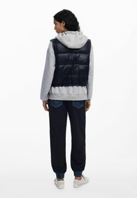 Mujer de espaldas vestida con un chaleco acolchado negro sobre una sudadera gris, pantalones jogger azul marino con bolsillos de mezclilla y zapatillas blancas.