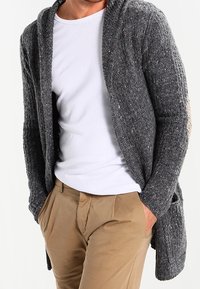 Šedý pletený cardigan s otevřeným předním dílem, dlouhými rukávy a texturovaným vzorem. Nositelný přes bílou košili a béžové kalhoty.