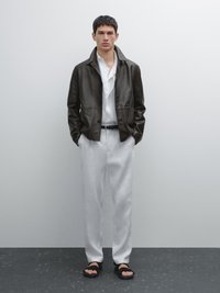 Dunkle Lederjacke mit geknöpftem Vorderteil, darunter ein weißes Hemd, locker sitzende weiße Hose und schwarze Sandalen. Strukturierten Hintergrund.