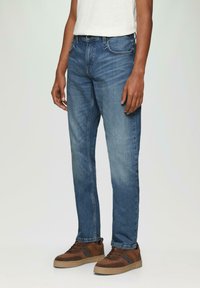 Donkerblauwe denim jeans met een rechte snit, vervaagde secties, vijf zakken en een knoopsluiting, gedragen met bruine en beige sneakers.