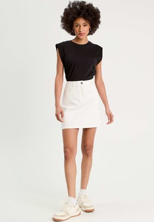 Camiseta negra de manga corta con hombreras, combinada con una minifalda de mezclilla blanca, que cuenta con un botón frontal y dos bolsillos laterales. Zapatillas blancas.