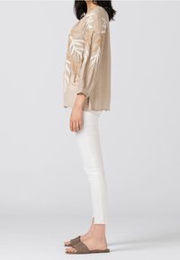 Femme portant un chemisier beige à manches longues avec une broderie de feuilles blanches et marron, un pantalon blanc ajusté et des sandales plates tressées marron, debout de profil.