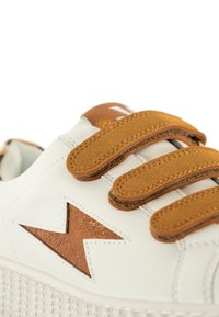 Sneaker blanc avec des sangles en suède marron à Velcro, un accent métallique marron en forme d'éclair, et une semelle en caoutchouc texturée.
