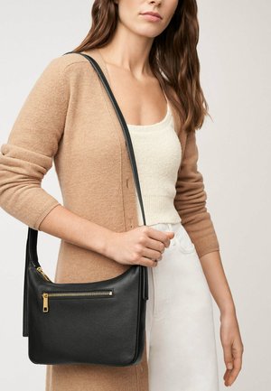 Sac en cuir noir à l'épaule avec une finition texturée, doté d'une poche zippée à l'avant et d'une bandoulière longue, porté de manière décontractée.