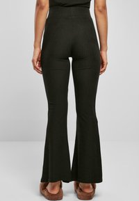 Pantalones acampanados de canalé verde oscuro con una alta cintura. La tela parece suave, con líneas verticales y una textura lisa. Se ven zapatillas de casa peludas.