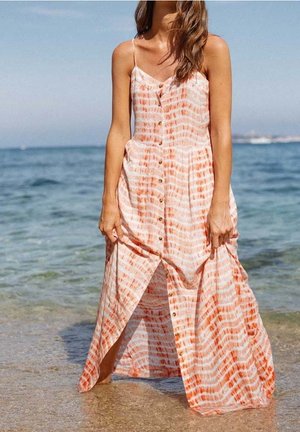 Vrouw in een oranje-wit gestreepte zomerse jurk met knopen, blootsvoets staand aan de rand van helder oceaanwater op een zonnige dag.