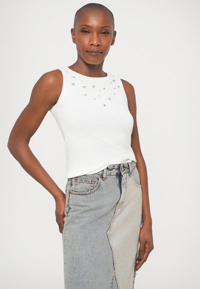 Fehér ujjatlan felső, kerek nyakkal, ezüst szegecsekkel díszítve a vállaknál. Kék és szürke árnyalatokkal ellátott hasított farmer szoknyával párosítva.