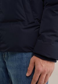 Navy blue puffer jacket with a smooth texture, angled stitch patterns, and a ribbed cuff. Denim jeans are visible underneath.