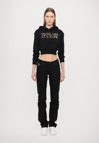 Zwarte cropped hoodie met "VERSACE JEANS COUTURE" in gouden letters, gecombineerd met zwarte jeans en witte sneakers. Model staat tegen een witte achtergrond.