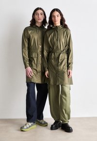 Olive green waterproof jackets with drawstring hoods, button closures, and waist belts, paired with dark jeans and cargo pants.
