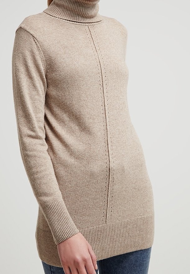 Pull à col roulé beige avec poignets et ourlet côtelés, présentant un motif vertical cousu au centre, porté avec un jean bleu.