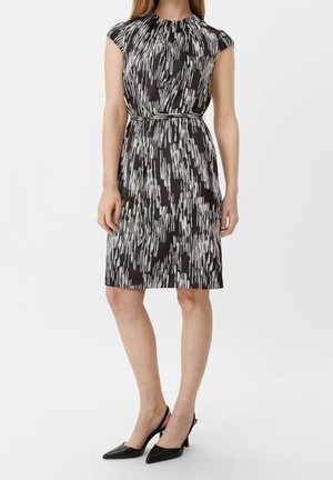 Kobieta ubrana w czarno-białą sukienkę bez rękawów z abstrakcyjnym wzorem do kolan, z cienkim paskiem i czarnymi, walenkami na obcasie z odkrytą piętą.