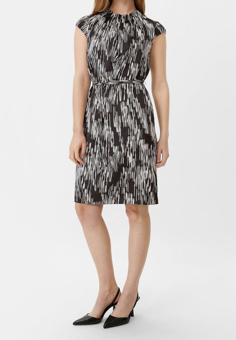Femme portant une robe sans manches mi-longue à motifs abstraits noir et blanc avec une ceinture fine et des escarpins slingback noirs à bout pointu.