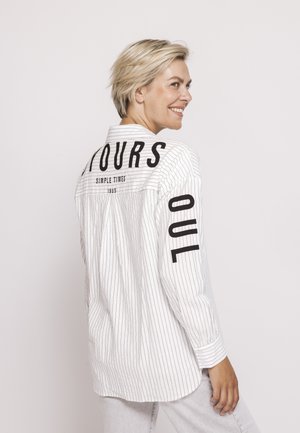 Smiling woman with short blonde hair wears white pinstripe shirt with black text on back and sleeve, standing against plain light background.