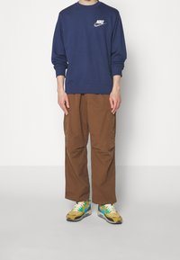 Sudadera azul marino con logo de Nike en blanco, pantalones cargo marrones con bolsillos laterales y zapatillas multicolor con detalles en amarillo, azul y negro.