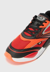 Red and black athletic shoe with mesh and synthetic materials, featuring a lace-up design, textured accents, and a white sole.