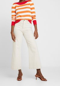 Pull rayé orange, blanc et rouge ; pantalon large crème taille haute ; talons bruns à brides. Design simple avec tissu texturé.