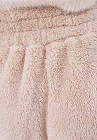 Zacht beige stof met een pluche, gestructureerd oppervlak dat gelaagde plooien vertoont. Materiaal lijkt op nepbont en benadrukt warmte en comfort.