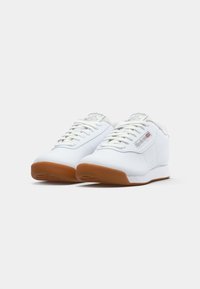 Par de ténis brancos Reebok Classic em pele com solas de borracha e atacadores brancos sobre fundo branco.