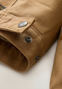 Manga de chaqueta de lona marrón con broches metálicos y un acabado texturizado. La tela presenta un suave tejido, añadiendo profundidad al diseño.