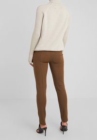 Vrouw in slimbruine broek, crèmekleurige ribgebreide coltrui en zwarte hoge hakken, staand tegen een effen grijze achtergrond.