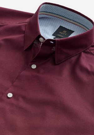 Chemise bordeaux avec un col boutonné, présentant une doublure intérieure bleue à motifs. Texture lisse, avec des boutons blancs et des détails de couture subtils.