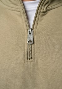 Lys beige hettegensere med glidelås, som har en sølvfarget glidelås, ribbede kanter og myk tekstur. Glidelåsen har et logo på trekkeren.
