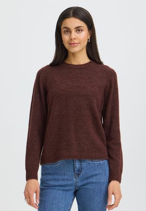 Femme aux longs cheveux foncés, portant un pull marron et un jean bleu, debout face à un fond clair et uni.