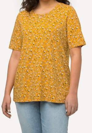 Kvinde iført en gul blomstret top med korte ærmer og lyseblå jeans, stående foran en ensfarvet hvid baggrund.