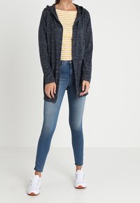 Mörkgrå huva cardigan med avslappnad passform, tillsammans med en gulrandig skjorta och blå jeans. Vita sneakers fulländar outfiten.