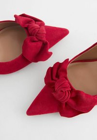 Czerwone zamszowe buty na szpilce z ostro zakończonym noskiem i węzełkowym detalem na przodzie. Gładka tekstura, beżowa wkładka, minimalistyczny design.