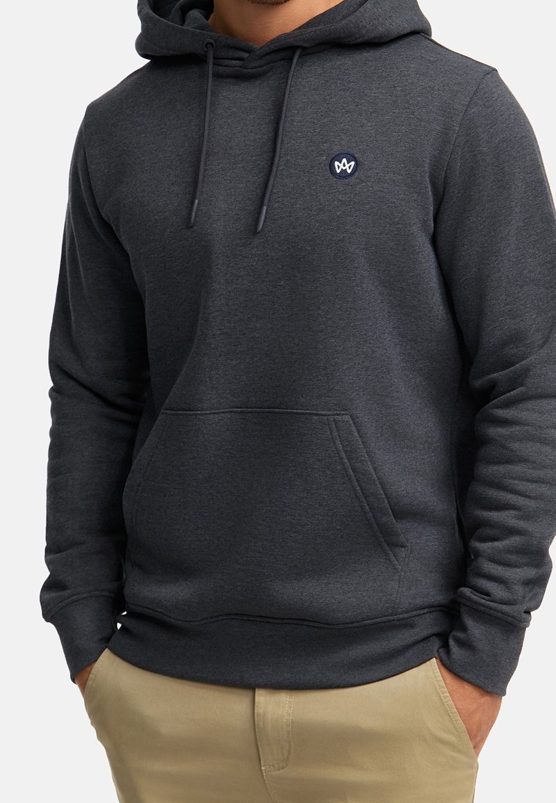 Homme portant un sweat à capuche gris foncé avec poche avant et petit logo sur la poitrine, mains dans les poches, pantalon beige visible.