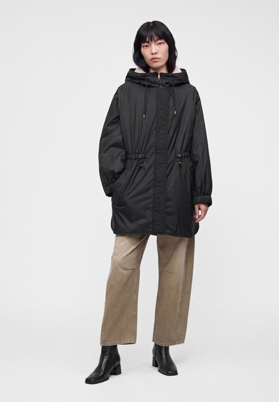 Femme portant un parka noir à capuche avec cordons, un pantalon beige à jambes larges et des bottines noires, debout devant un fond uni.