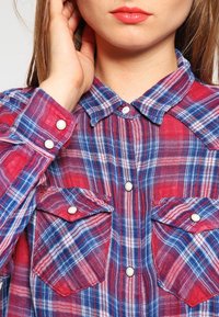 Vrouw in een rood-blauw geruit overhemd met knoopsluitingen en zakken met drukknopen, die haar haar bij het oor aanraakt.