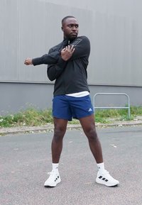 Černá sportovní bunda, modré šortky a bílé tenisky. Bunda má zip a otvory pro palce; šortky mají logo Adidas.