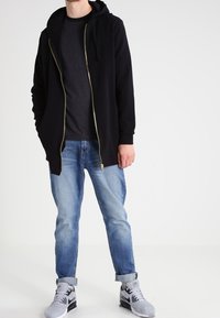 Svart zip-up hoodie med guld dragkedja, lager på ett mörkt t-shirt. Kombinerad med ljusblåjeans och grå träningsskor.
