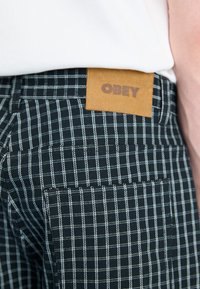 Mustristest mustade ja valgete kantidega püksid, millel on pruun nahkbool, millel on kirjas "OBEY", valged õmblused ja tekstureeritud kangas.