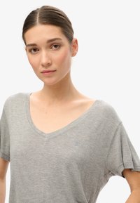 Camiseta gris de manga corta con cuello en V, hecha de una tela suave, que presenta un logo bordado sutilmente en el pecho. Diseño simple y de corte relajado.