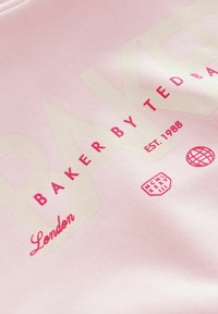 Gaiši rozā džemperis ar lieliem krēmkrāsas burtiem "BAKER BY TED BAKER", ar sarkaniem akcentiem un grafiskām detaļām, tostarp emblēmu un datumu.