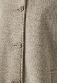 Beige ullfrakk med herringbone-mønster, med fire store brune knapper, en strukturert krage og en myk tekstur.