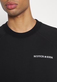 Sudadera negra con cuello acanalado y mangas raglán. Presenta el logotipo blanco "SCOTCH & SODA" en el lado izquierdo del pecho. Tejido suave y texturizado.