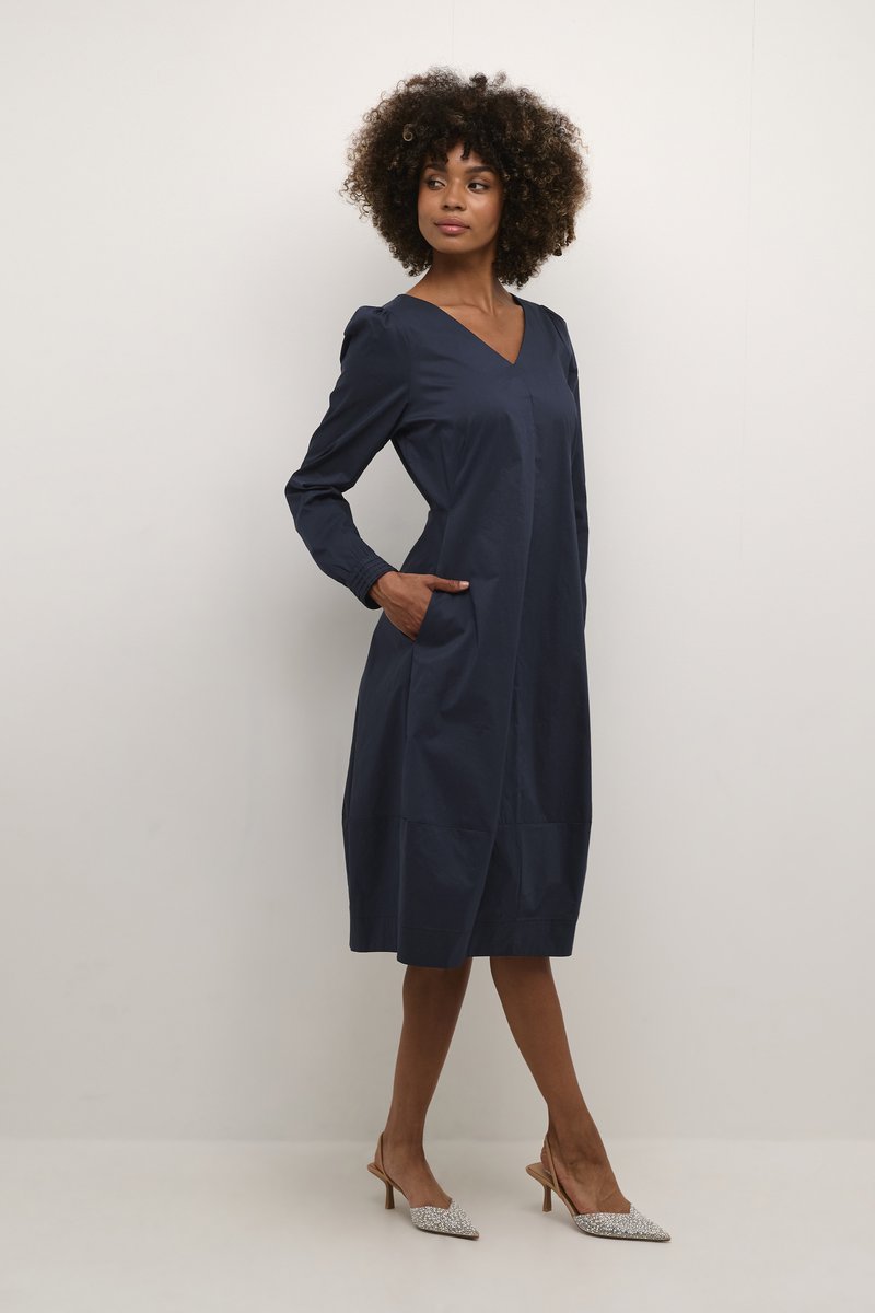 Tumesinine, pikkade varrukatega kleit V-kaelusega, küljetaskudega ja kogutud õlgadega. Kantud sädelevate kontsadega. Sile tekstuur.