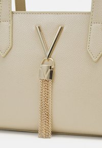 Beige laukku, jossa on tekstuuri, kultainen "V"-embleemi ja ketjutupsu, jossa on riippulukko. Olkahihnat, joissa on kulmikkaat suunnitteluakcentit.
