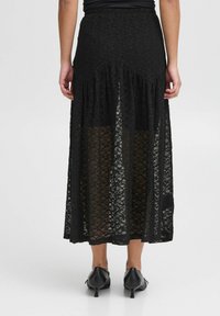 Černá krajková maxi sukně s vrstveným designem, obsahující průhledné panely a nařasený pás. Textura látky je složitá a lehká.